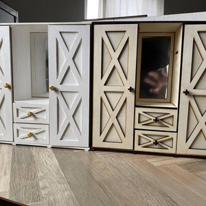 May include: Two miniature wooden wardrobes featuring X-patterned doors, two stacked drawers, and a central mirror. One wardrobe is painted solid white with gold-toned knobs, while the other is natural light wood with dark brown edges and dark knobs.