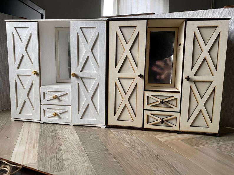 May include: Two miniature wooden wardrobes featuring X-patterned doors, two stacked drawers, and a central mirror. One wardrobe is painted solid white with gold-toned knobs, while the other is natural light wood with dark brown edges and dark knobs.