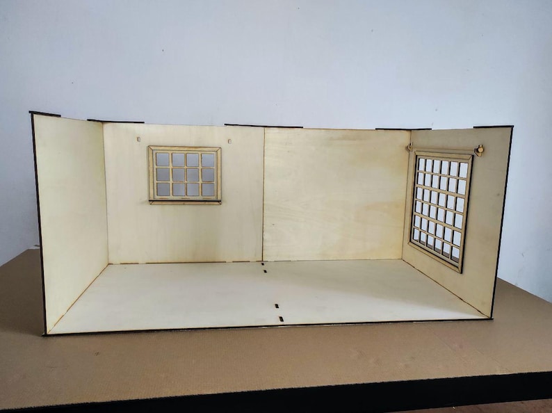 May include: A light-colored wooden dollhouse room structure featuring three walls and a floor. The back wall includes a small square window with a grid pattern, while the right side wall features a larger rectangular window with a grid design.
