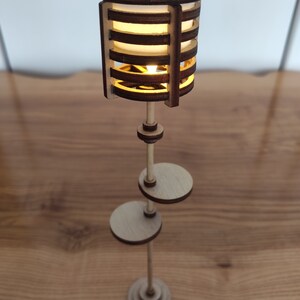 May include: A wooden floor lamp with a cylindrical shade made of thin wooden slats. The lamp has a warm, white light and a simple, modern design.