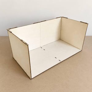 May include: A rectangular open-top box constructed from light-colored plywood panels. The structure features visible interlocking joints at the corners and edges, with dark brown laser-cut edges contrasting against the pale wood surfaces.