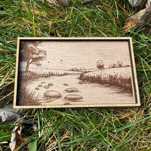 May include: A wooden plaque with a laser-engraved image of a river scene. The scene features a winding river with rocks in the water, a tree on the left side, and a grassy field on the right side.