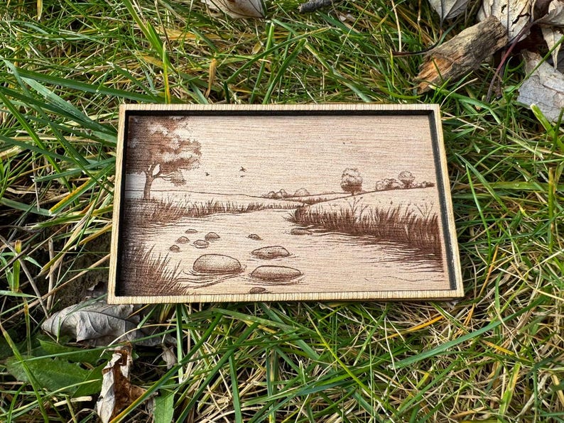 May include: A wooden plaque with a laser-engraved image of a river scene. The scene features a winding river with rocks in the water, a tree on the left side, and a grassy field on the right side.