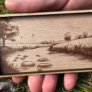 May include: A wood-burned image of a stream with rocks in the water, surrounded by trees and grass. The image is framed in a wooden frame.