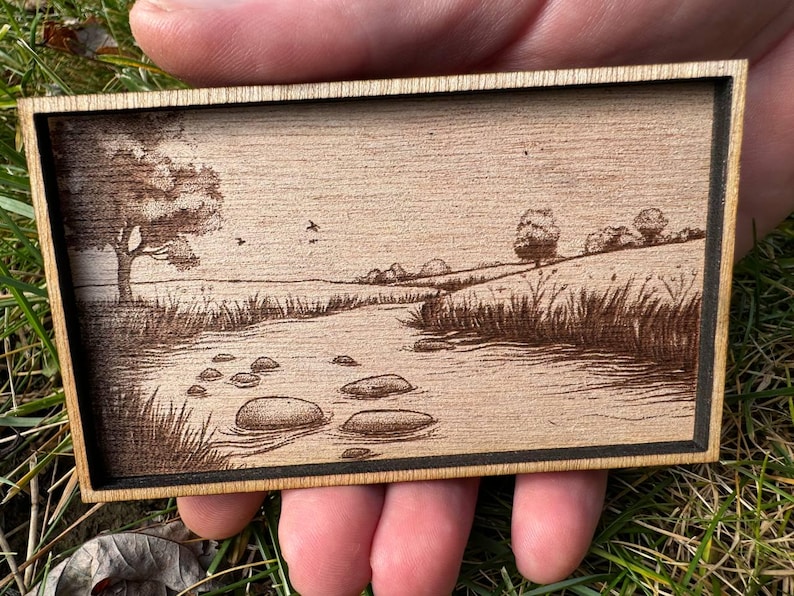 May include: A wood-burned image of a stream with rocks in the water, surrounded by trees and grass. The image is framed in a wooden frame.