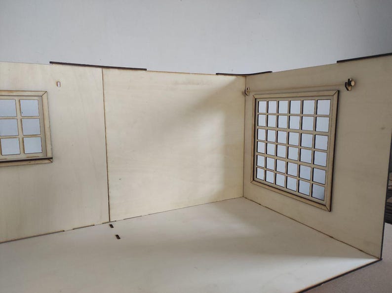 May include: A light-colored wooden dollhouse room structure featuring two walls and a floor. The left wall has a small window with six panes, while the right wall contains a larger window with a grid of thirty-five square panes. The wood is smooth.