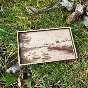 May include: A wooden framed artwork with a brown and white landscape scene of a river with rocks and trees. The artwork is on a green grassy surface.