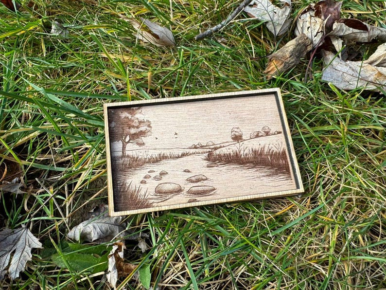 May include: A wooden framed artwork with a brown and white landscape scene of a river with rocks and trees. The artwork is on a green grassy surface.