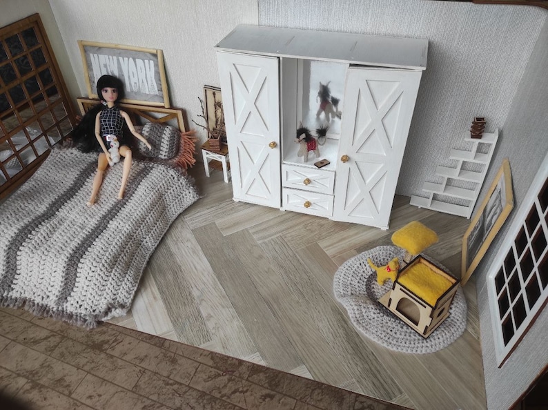 May include: A miniature dollhouse bedroom set featuring a bed with a gray and white striped crocheted blanket, a white wooden wardrobe with X-patterned doors, a small side table, a white tiered shelf, a round gray rug, and a yellow cat-themed pet house.