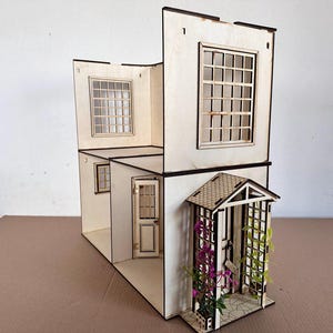 May include: A two-story wooden dollhouse structure with light-colored walls, multiple grid-patterned windows, and a front door. The exterior features a small porch with a peaked roof, a trellis, and artificial purple and green climbing plants.