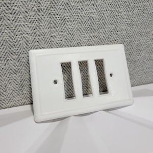 May include: A white, rectangular wall plate with three rectangular cutouts. The plate has a raised border and two small holes for mounting. The background is a gray and white herringbone pattern.