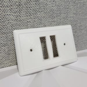 Pode incluir: Uma placa de interruptor de luz branca e retangular com duas aberturas retangulares para interruptores. A placa tem uma borda elevada e dois orifícios para parafusos. O item é feito de um material liso e sólido.
