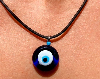Pode incluir: Um colar com um amuleto azul de olho turco com um cordão de couro preto. O amuleto é redondo e tem um olho branco com uma pupila azul.