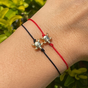 Puede incluir: Dos pulseras de la amistad con dijes de tortuga dorada. Una pulsera es de hilo rojo y la otra de hilo negro.