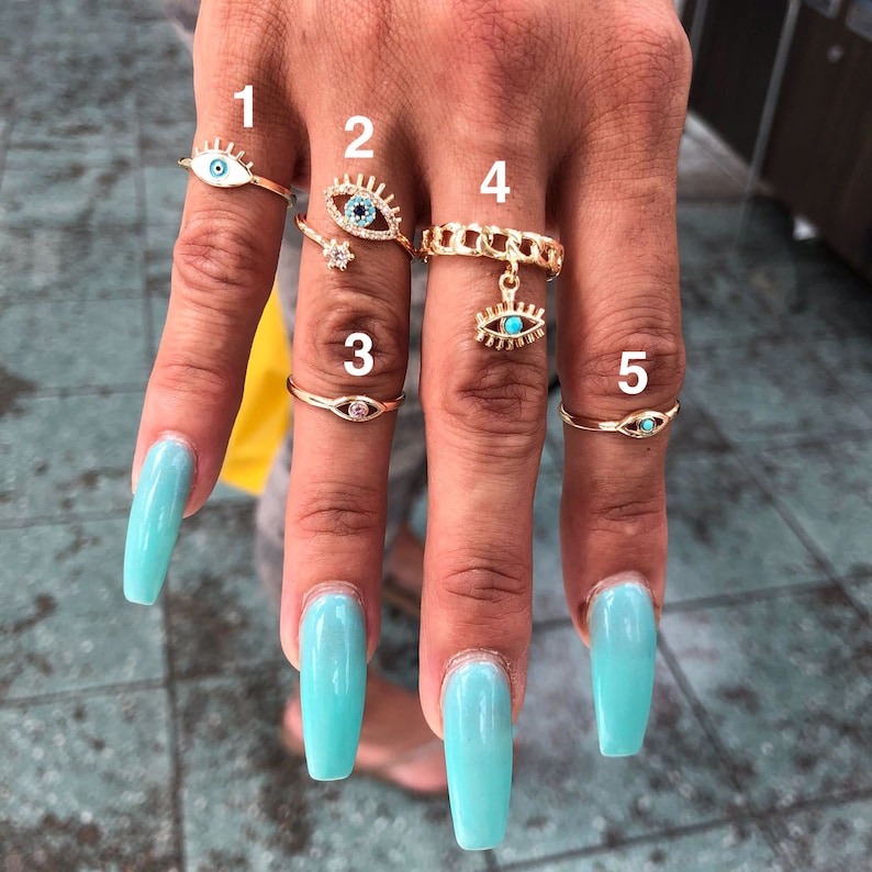May include: Five gold rings with blue evil eye charms. The rings are stacked on a hand with light blue nail polish.