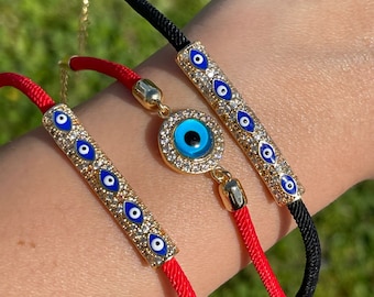 Può includere: Tre braccialetti dorati con ciondoli a forma di occhio blu. Due braccialetti hanno cordini rossi e uno ha un cordino nero. I ciondoli a forma di occhio sono circondati da cristalli trasparenti. I braccialetti sono mostrati su un polso.