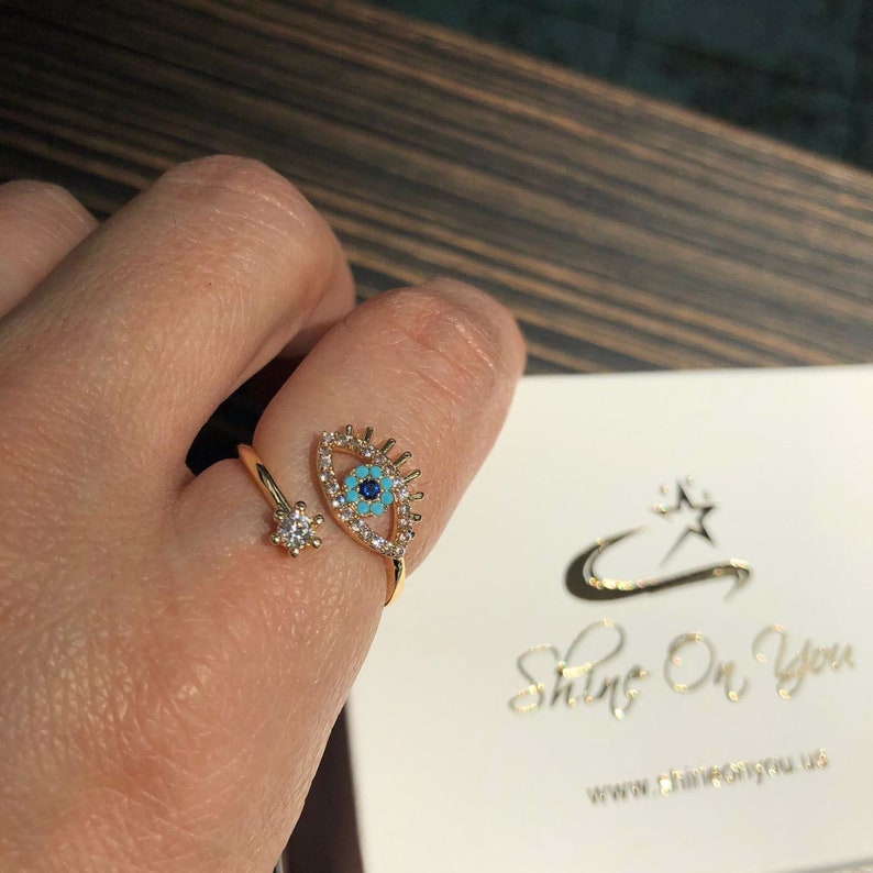 May include: Gold ring with a blue and turquoise evil eye design and a small diamond accent. The ring is adjustable and can be worn on any finger.