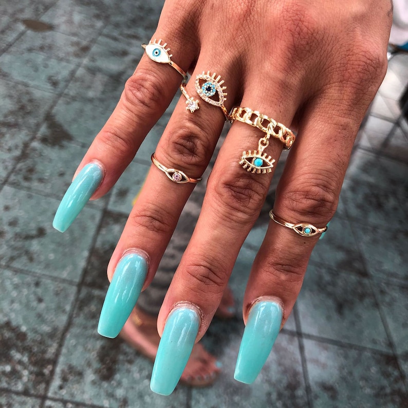 May include: A hand with light blue nail polish is wearing five gold rings with blue evil eye charms. The rings are on the index, middle, ring, and pinky fingers.
