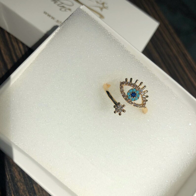 May include: Gold ring with a blue evil eye design and clear rhinestones. The ring is open and adjustable.