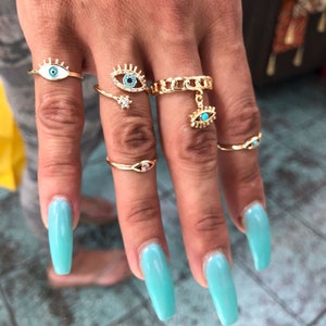 May include: A set of five gold rings with blue and white evil eye charms. The rings are worn on the fingers of a hand with long, light blue nail polish.