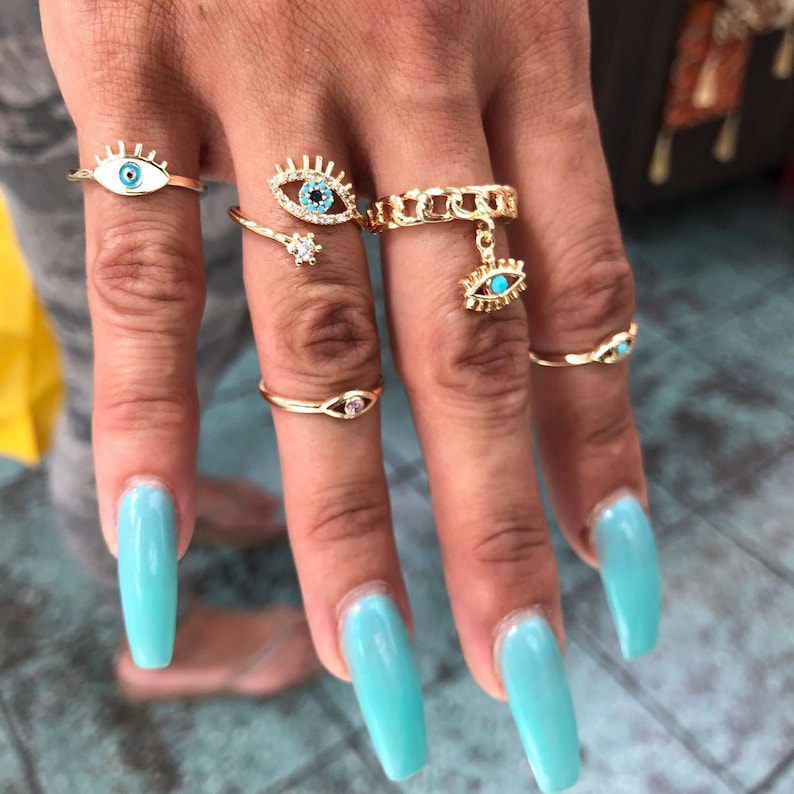 May include: A set of five gold rings with blue and white evil eye charms. The rings are worn on the fingers of a hand with long, light blue nail polish.