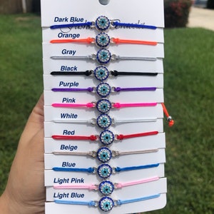 May include: A set of 12 evil eye bracelets in various colors, including dark blue, orange, gray, black, purple, pink, white, red, beige, blue, light pink, and light blue. Each bracelet features a silver charm with a blue evil eye design.