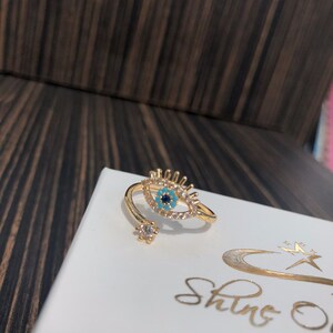 May include: Gold ring with a blue and turquoise evil eye design, surrounded by clear rhinestones. The ring is open and adjustable.