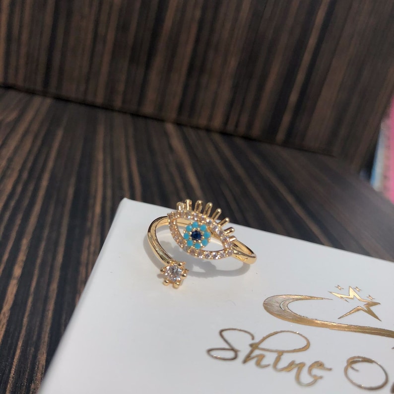 May include: Gold ring with a blue and turquoise evil eye design, surrounded by clear rhinestones. The ring is open and adjustable.