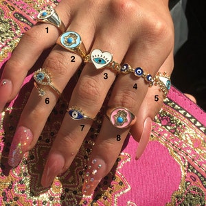 May include: A set of eight gold rings featuring blue and turquoise evil eye charms and hamsa hand charms. The rings are worn on a hand with long, manicured nails.