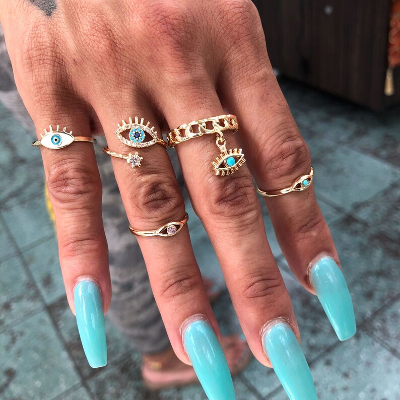 May include: A set of five gold rings with blue evil eye charms. The rings are worn on the fingers of a hand with light blue nail polish.