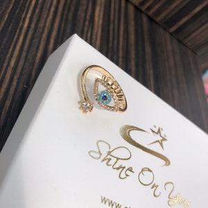 May include: Gold ring with a blue and turquoise evil eye design. The ring is set with clear rhinestones and has a small flower-shaped accent.