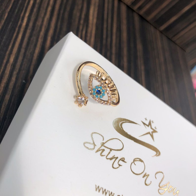May include: Gold ring with a blue and turquoise evil eye design. The ring is set with clear rhinestones and has a small flower-shaped accent.