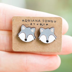 May include: A pair of gray and white fox-shaped earrings on a small brown card. The earrings have a cartoon-like design with a white muzzle and black nose. The card has the text "ADRIANA TOMEU STUDIO" printed on it.