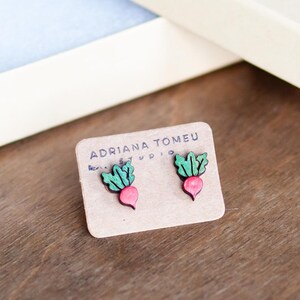 Könnte beinhalten: Ein Paar Holzohrringe in Form von Radieschen mit grünen Blättern. Die Ohrringe werden auf einer kleinen Karte mit dem Text "Adriana Tomeu Studio" präsentiert.