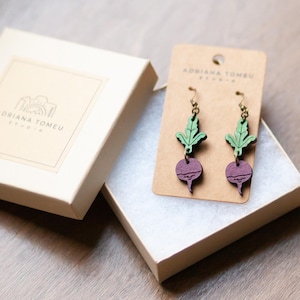 May include: A pair of wooden earrings shaped like radishes with green tops. The earrings are packaged in a white box with the text "Adriana Tomeu Studio" on the lid.