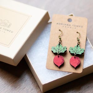 May include: A pair of wooden earrings shaped like radishes with green leaves and red bulbs, displayed on a cardboard tag inside a white box. The earrings have gold-colored hooks. The box has the text "ADRIANA TOMEU STUDIO".