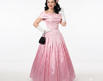 May include: A floor-length, off-the-shoulder pink satin gown with a fitted bodice and a full, pleated skirt. The ensemble includes white elbow-length gloves and a small black handbag with a silver frame, worn with a pink hair flower accessory.