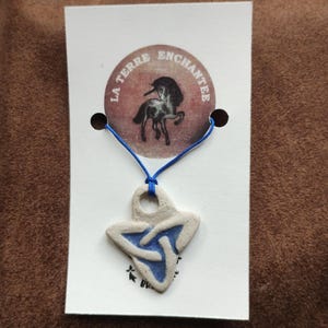 Peut inclure: Un pendentif en céramique crème avec un motif de nœud celtique bleu, suspendu à un cordon bleu. Le pendentif est attaché à une carte blanche avec le texte "LA TERRE ENCHANTEE" et une illustration de licorne.