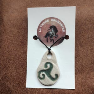 Peut inclure: Un pendentif en céramique blanche avec un nœud celtique vert, suspendu à un cordon noir. Le pendentif est sur une carte blanche avec une image en noir et blanc d'un cheval et le texte "La Terre Enchantee".