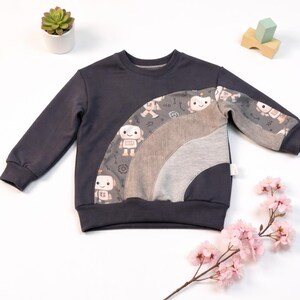Könnte beinhalten: Dunkelgraues Kinder-Sweatshirt mit einem Regenbogen-Design mit Roboter-Illustrationen. Der Regenbogen besteht aus grauem und beigem Stoff. Das langärmlige Oberteil hat einen Rundhalsausschnitt und ist für Freizeitkleidung geeignet.