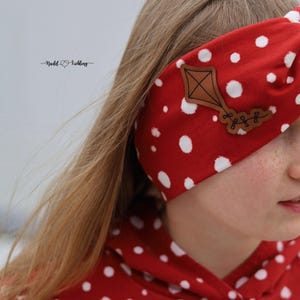 Könnte beinhalten: Ein rotes Stirnband mit weißen Punkten und einem braunen Drachen-Applikation. Das Stirnband ist Teil eines passenden Sets, sichtbar mit einem roten Kapuzenpullover mit Punkten. Der Text "Naddl & Liebling" ist sichtbar.