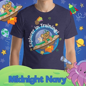 May include: A navy blue t-shirt with a cartoon bear and rabbit in a spaceship. The text "Explorer in Training!" is written in a circle around the spaceship. The shirt is on a model and the background is blue with stars and other space-themed graphics.