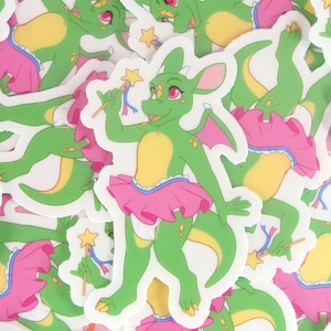May include: A pile of green and pink dragon stickers with a star wand. The dragons are wearing pink tutus and have yellow accents.