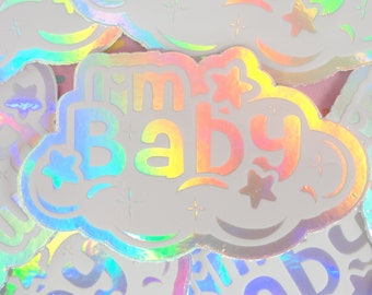 May include: A holographic sticker with the text "I'm Baby" in a rainbow color scheme. The sticker is shaped like a cloud and has stars around the text.
