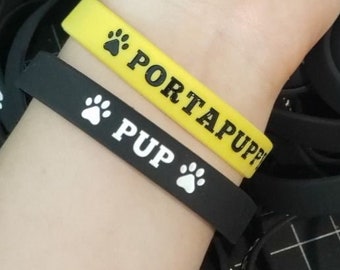 May include: A collection of silicone wristbands in black and yellow. The black bands feature the white text PUP and a paw print icon. The yellow band features the black text PORTAPUPPY and a paw print icon. These flexible rubber bracelets are designed for comfortable wear.