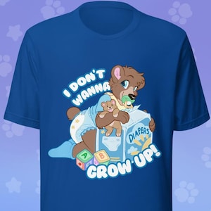 May include: A royal blue short-sleeve t-shirt featuring a graphic of a brown bear character wearing a diaper and holding a pacifier. The bear sits on a box labeled DIAPERS with the text I DON'T WANNA GROW UP! and alphabet blocks.