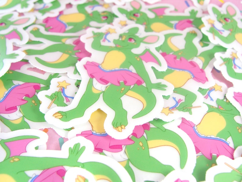 May include: A pile of stickers featuring a green dragon wearing a pink tutu and holding a magic wand. The dragon has yellow accents and a pink bow on its head.
