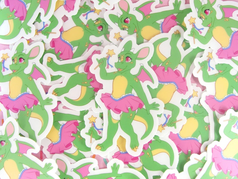 May include: A pile of green and pink dragon stickers with a star wand. The dragons are wearing pink tutus and have yellow accents.