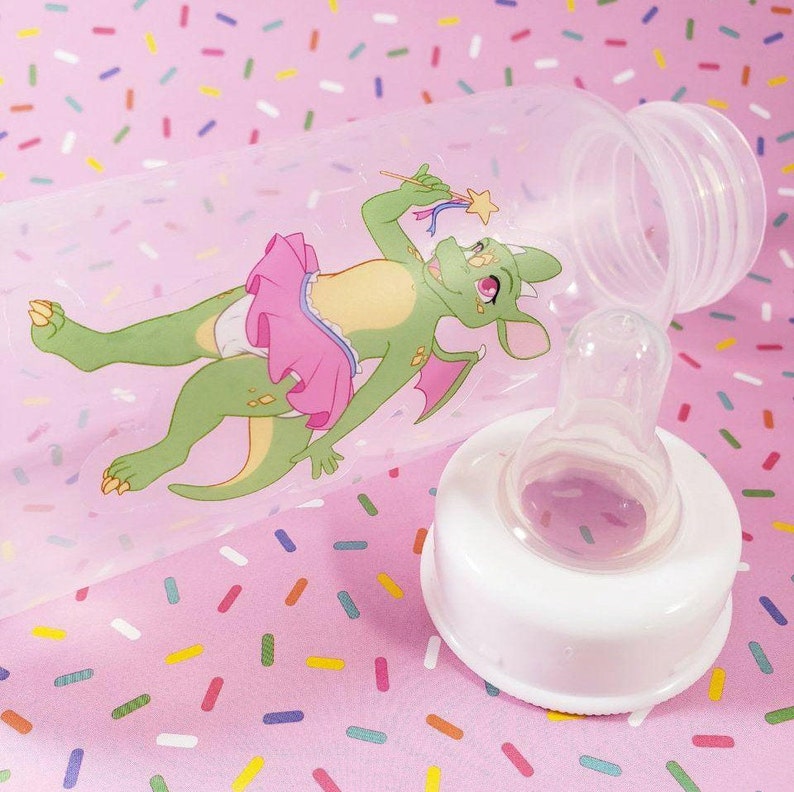 May include: A clear plastic baby bottle with a white nipple and a green dragon wearing a pink tutu and holding a wand. The bottle is on a pink background with colorful sprinkles.