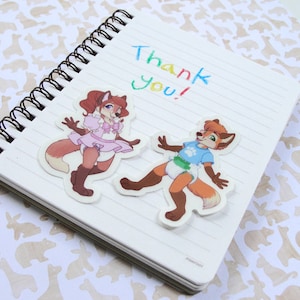 May include: A white notebook with a spiral binding and the words "Thank You!" written in colorful marker. Two cartoon fox stickers are attached to the notebook. One fox is wearing a pink dress and the other is wearing a blue shirt and a diaper.